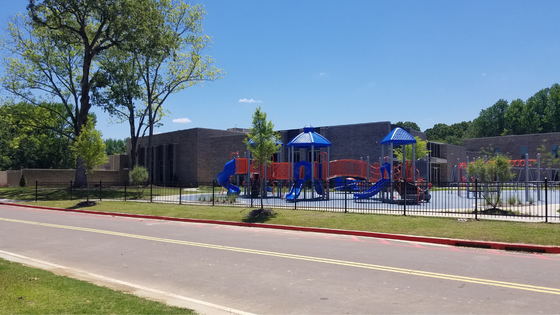 Playground with blue and red equipment, behind a black fence, in front of a brick building on a sunny day.