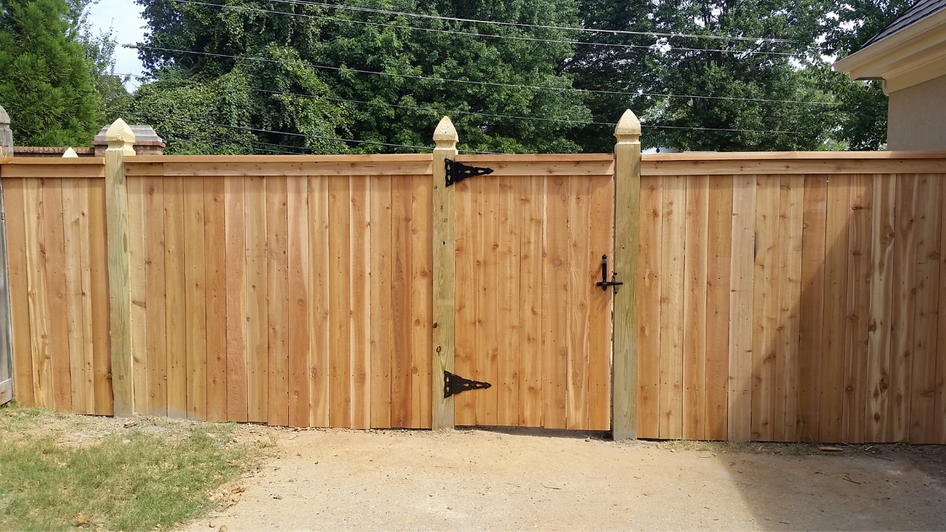 Wooden fence with gate, light brown, set on gravel, with trees in the background.