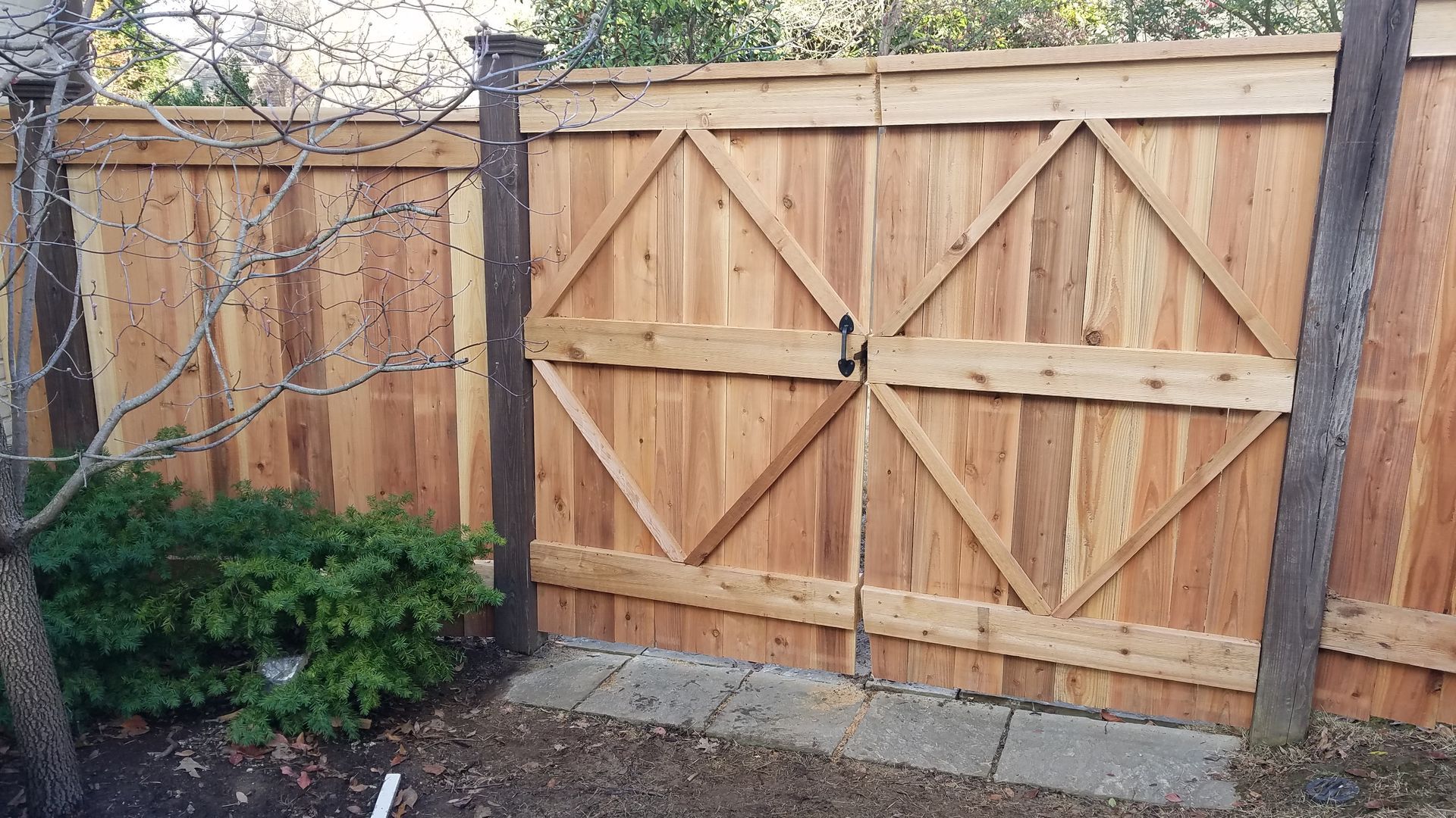 A wooden double gate with diamond-pattern bracing in a fenced yard, flanked by dark posts near a small bush.
