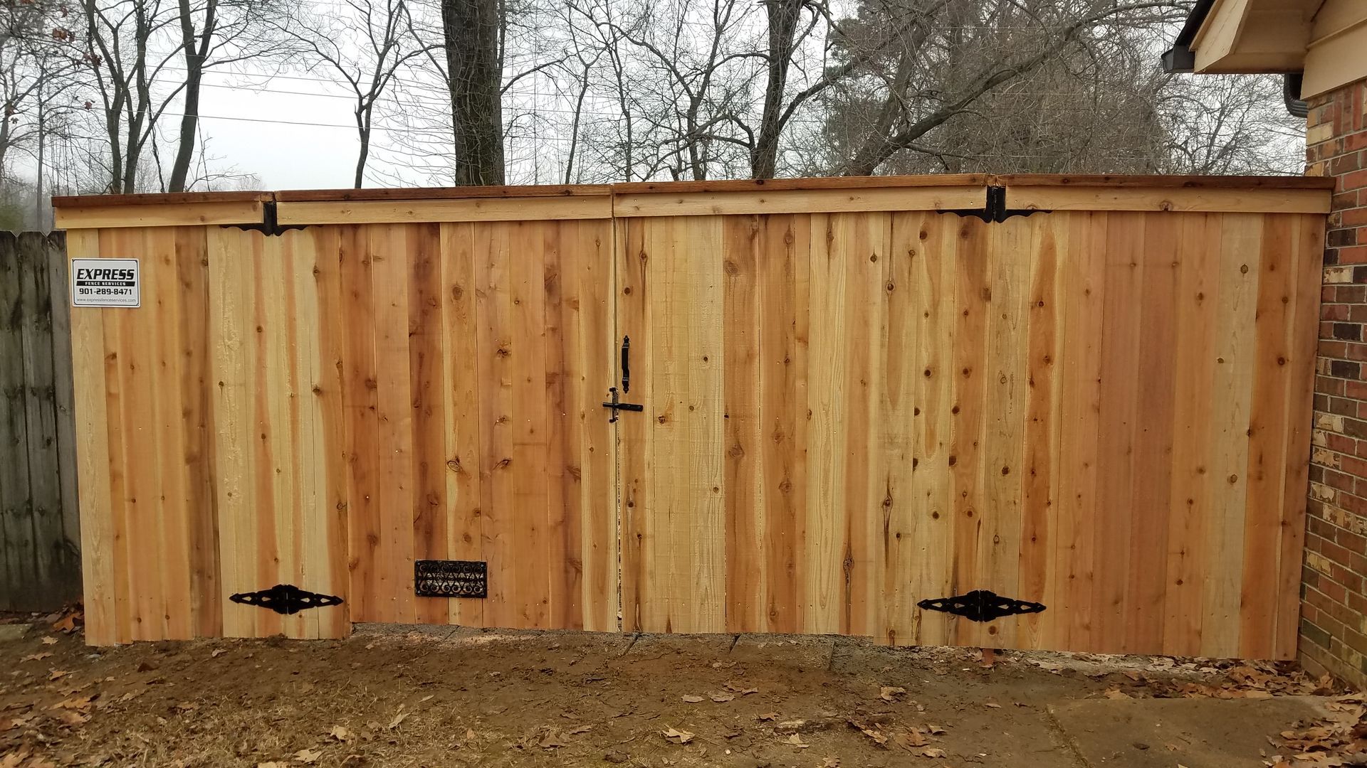 A wide, light-colored wooden double gate installed in a backyard fence with black metal hinges and a central latch.