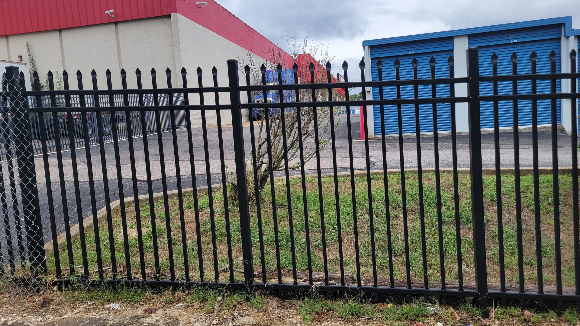 A black metal picket fence in front of a green patch of grass, with industrial buildings in the background.