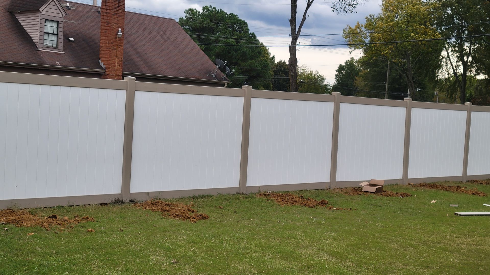 A white vinyl privacy fence with tan trim posts, standing in a residential lawn in front of a house.