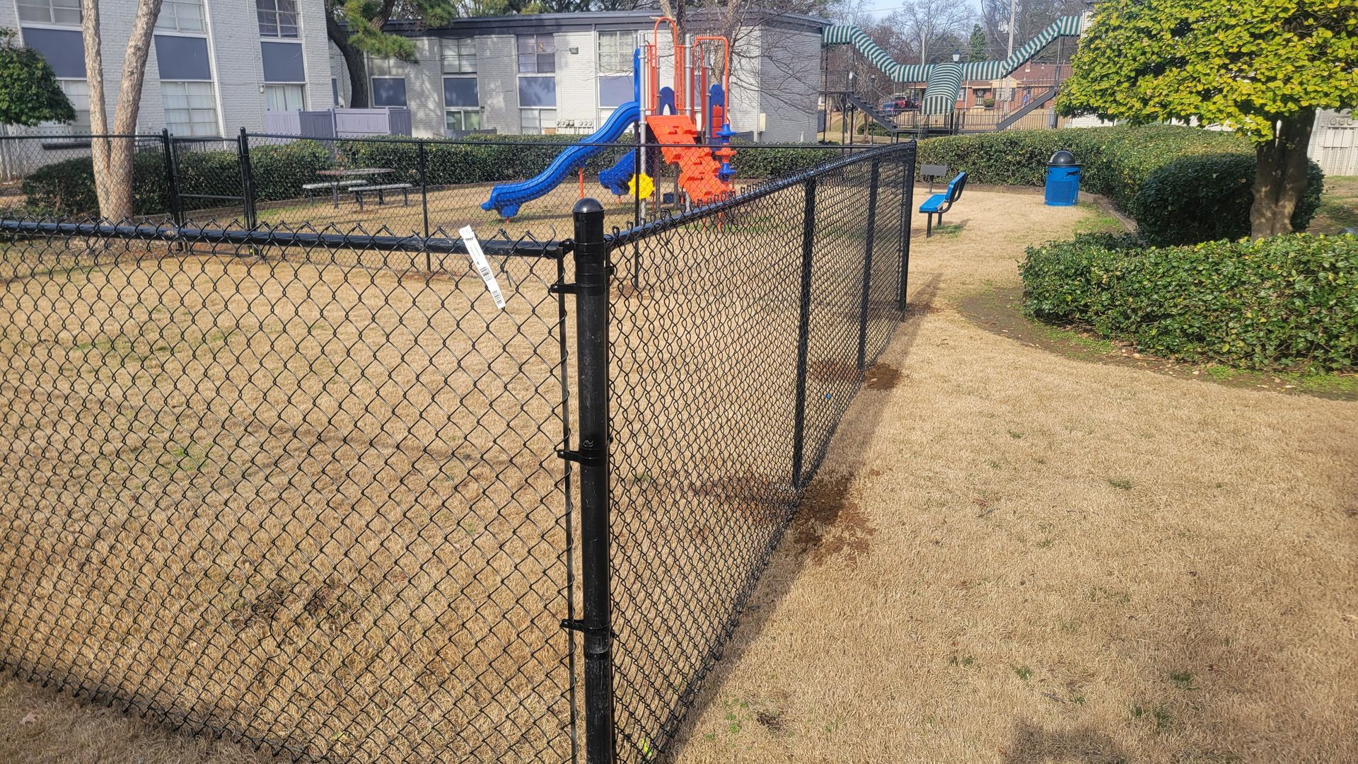A black chain-link fenced playground with a blue slide and an orange climbing structure on a dry, grassy field.