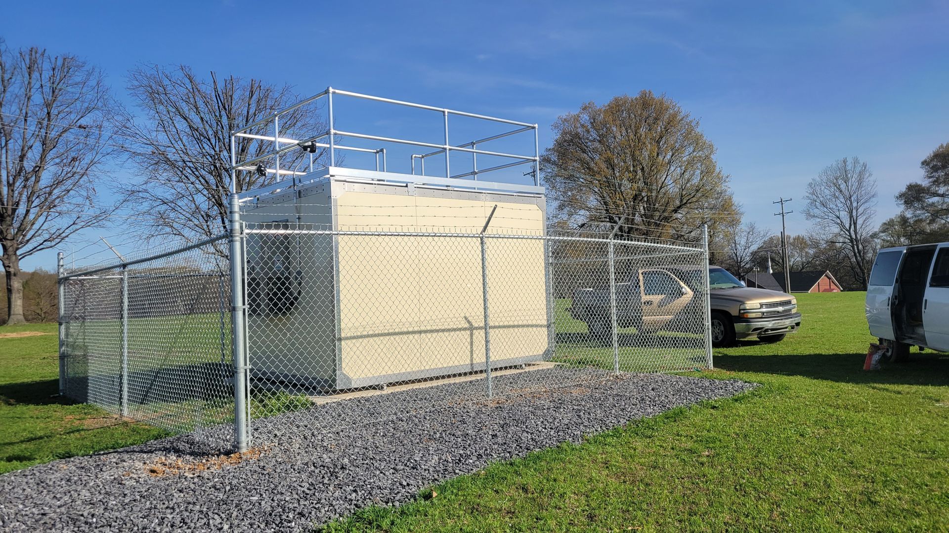 A fenced, beige utility building sits on a gravel pad in a grassy field under a clear blue sky.