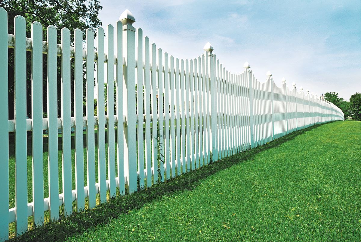 White picket fence curves along a green grassy yard under a bright sky.
