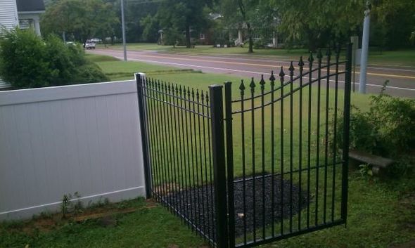 Black metal gate set into a white vinyl fence, opening onto a grassy area beside a street.