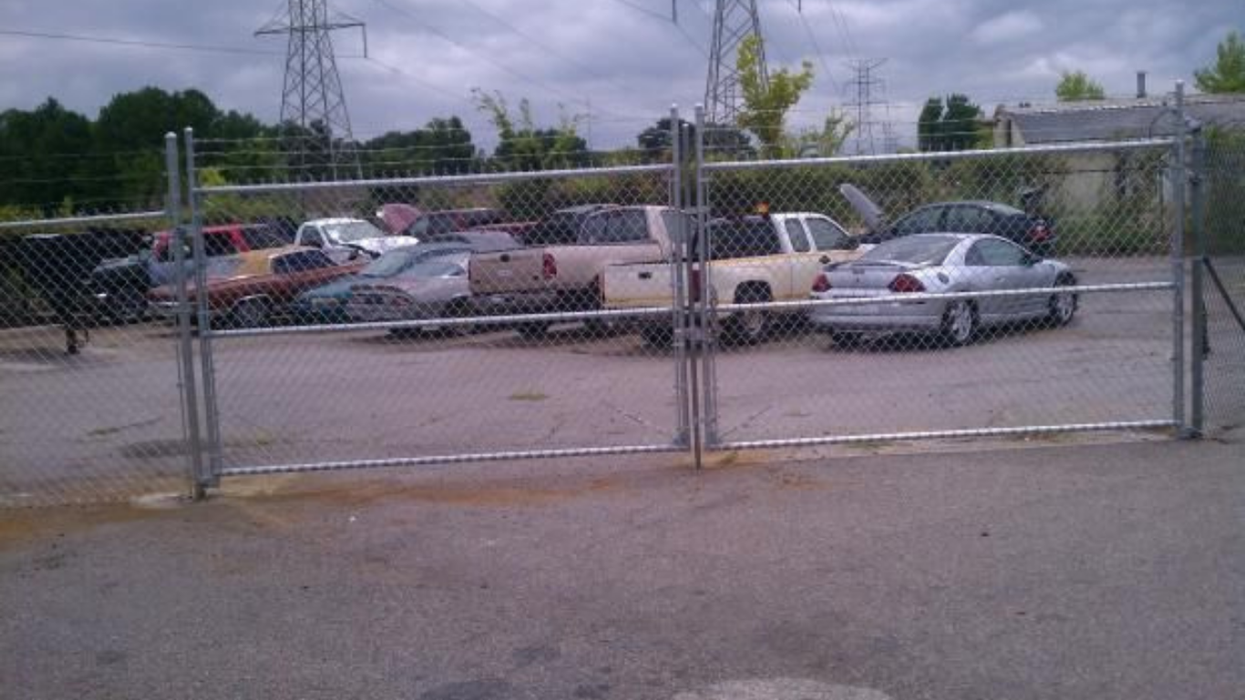 Chain-link fence in front of a parking lot with several vehicles. Power lines and cloudy sky in background.
