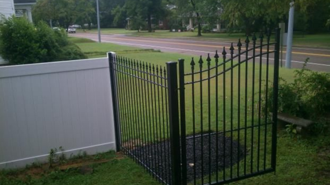 Black iron gate in front of a white vinyl fence with a road and green grass in the background.