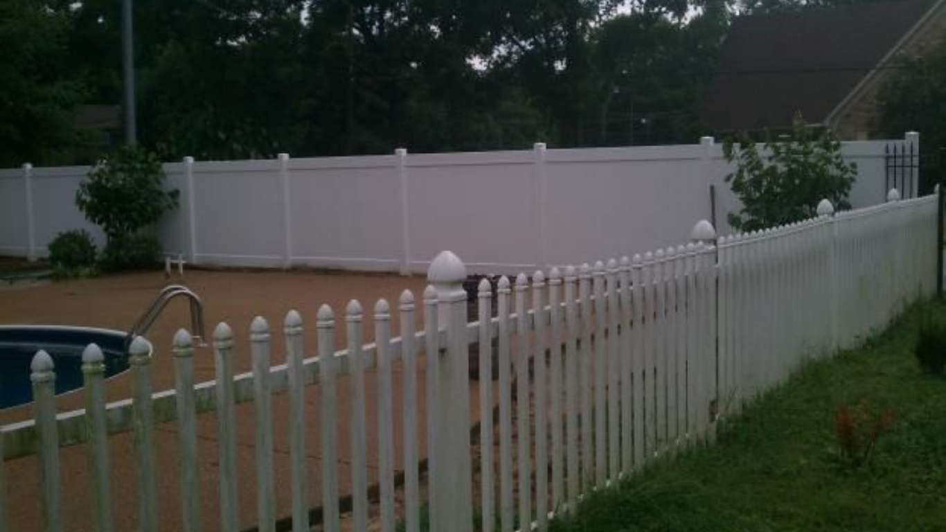 White picket fence next to a pool and a taller white fence in the background, trees in the distance.