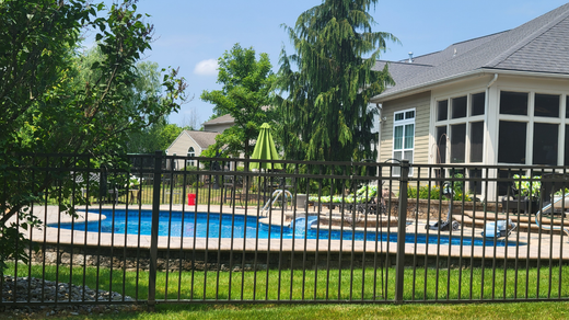 Black fenced-in backyard pool with green grass, trees, and a house with a screened-in porch on a sunny day.