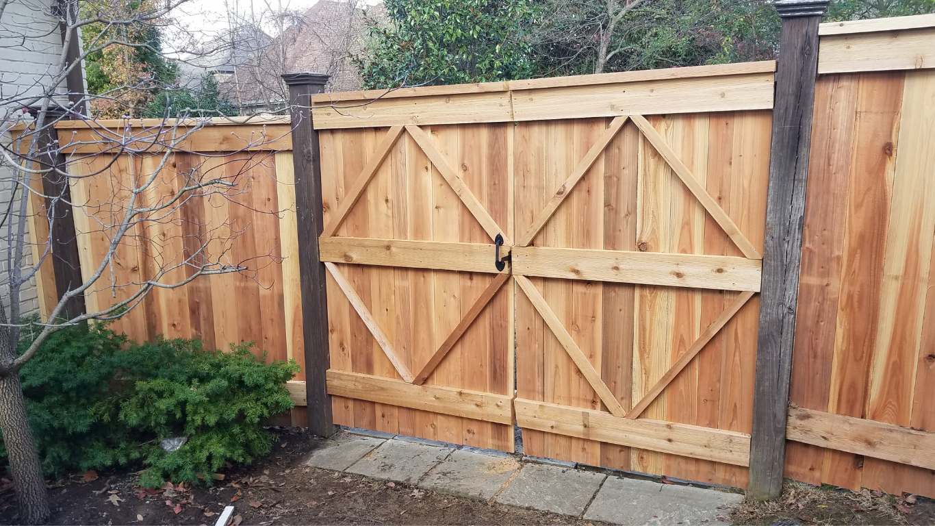 Wooden gate in a cedar fence with diamond-shaped accents; green shrubs and a paved walkway visible.