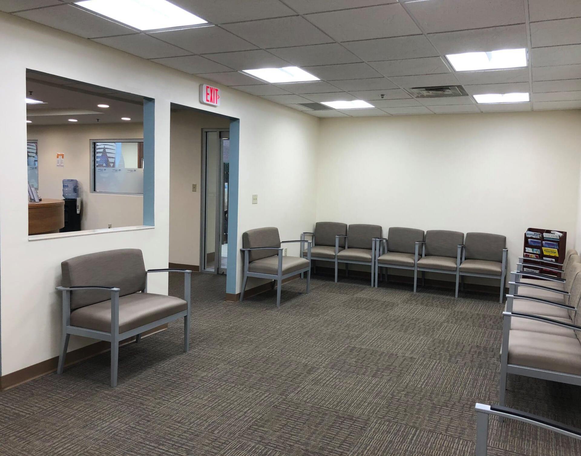 Waiting room with several gray chairs, exit sign, and a window.