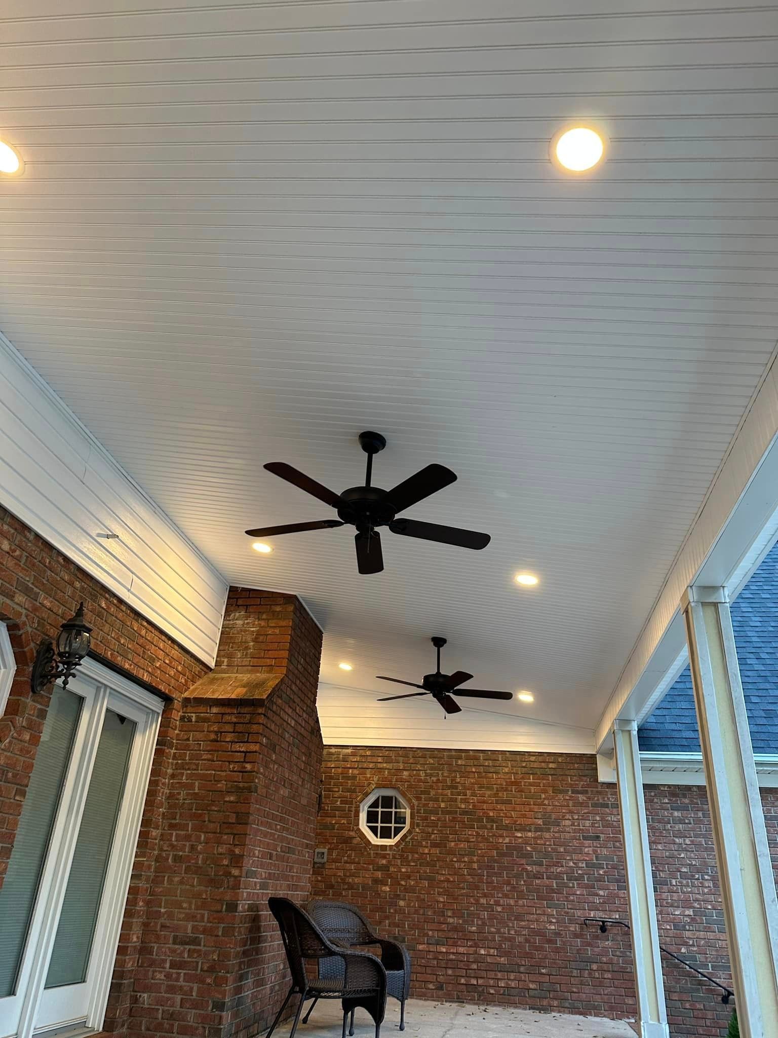 Patio ceiling with two black ceiling fans and recessed lights. Red brick wall and outdoor furniture.