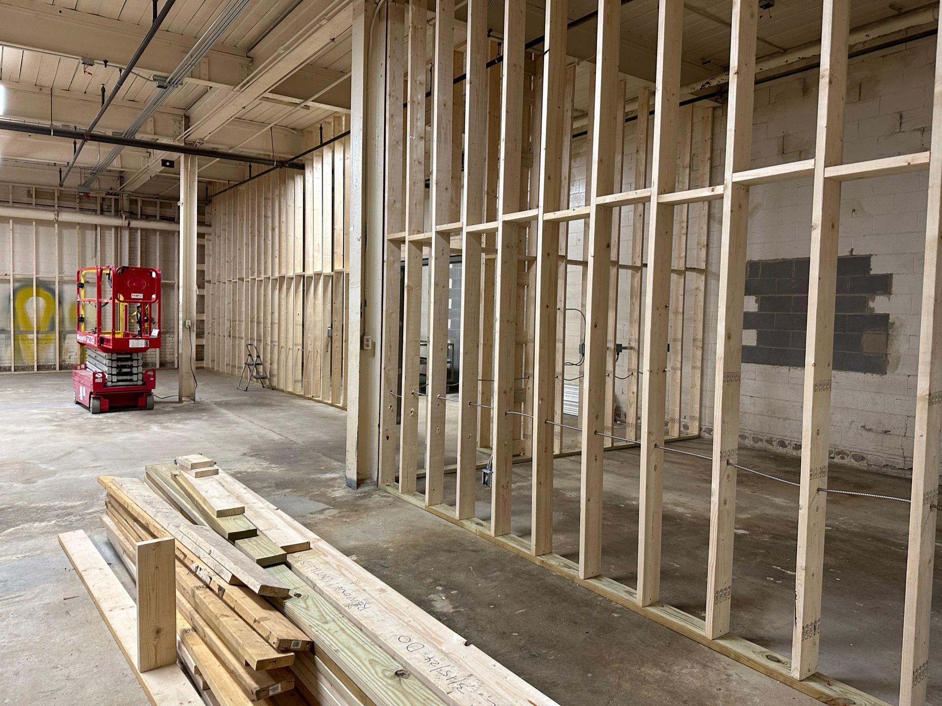 Interior view of a construction site. Wooden studs frame new walls. A red lift and lumber are visible.