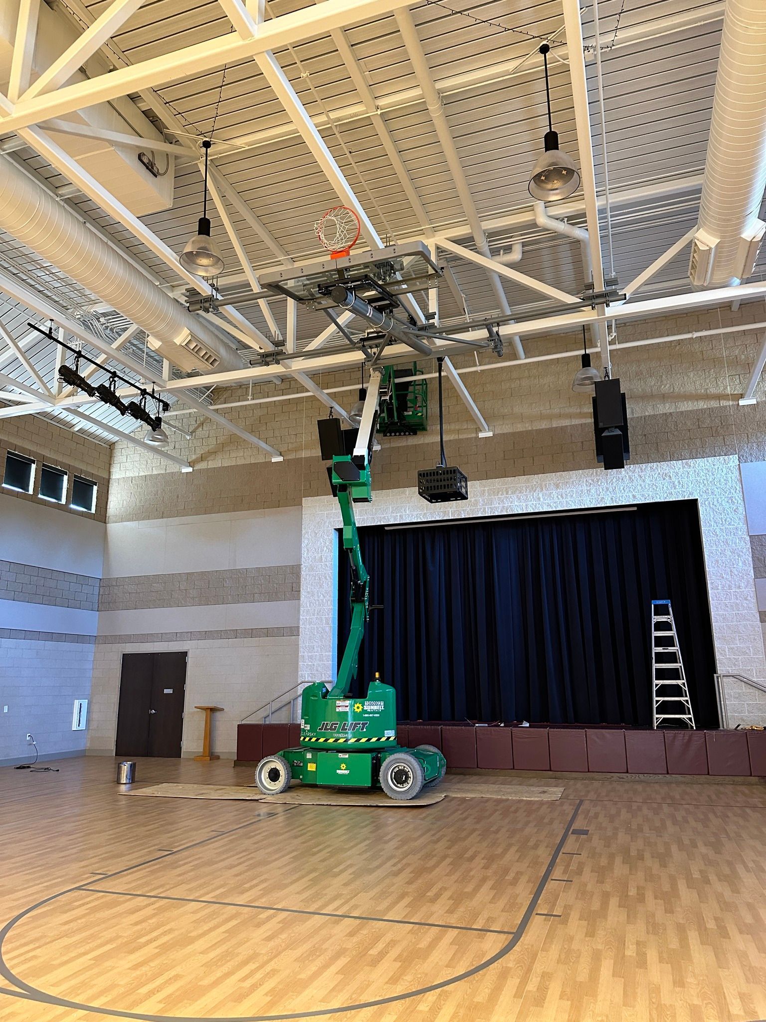 A green lift supporting speakers and equipment inside a gymnasium. A stage with a black curtain is in the background.