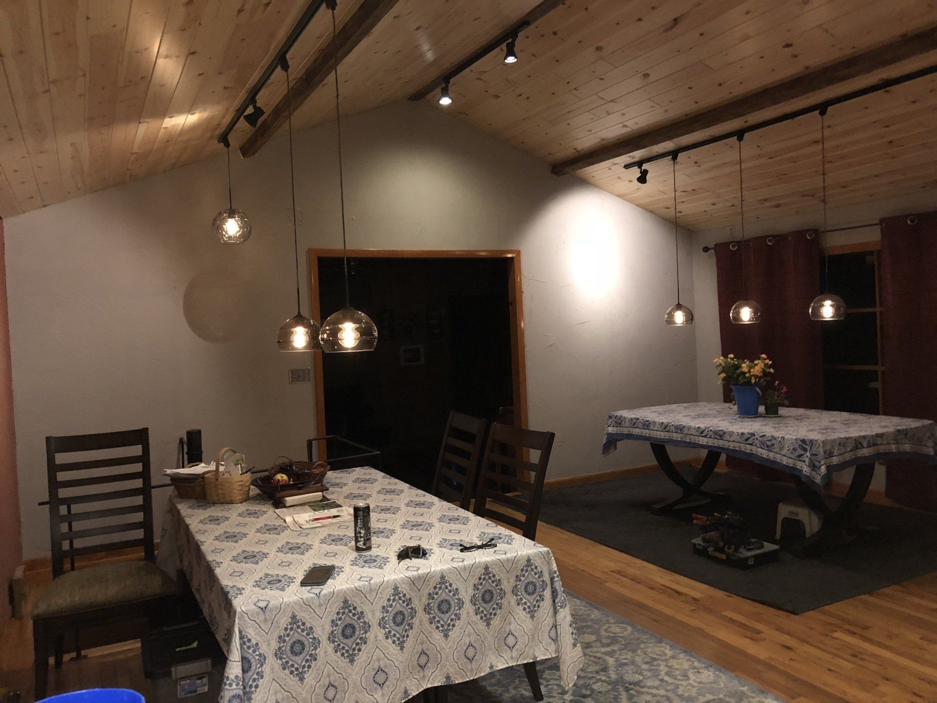Dining room with two tables covered in patterned tablecloths, pendant lights, and wooden ceiling.