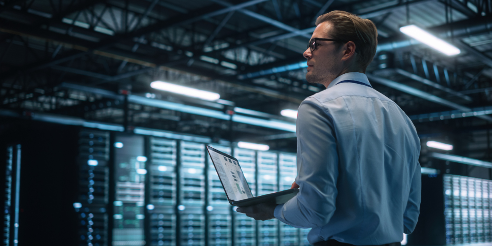 A person in a light blue shirt holds a laptop while standing in a dimly lit, professional server room.