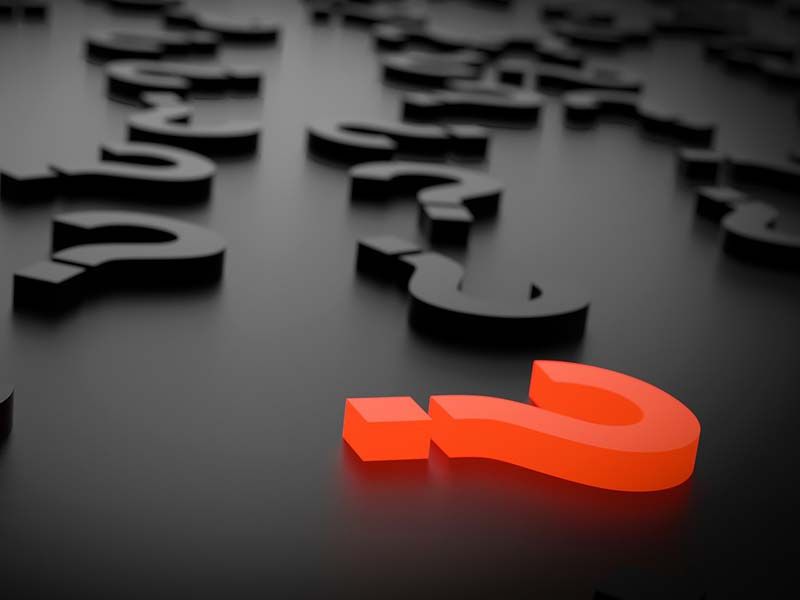 A vibrant orange question mark stands out on a dark surface among many blurred black question marks.