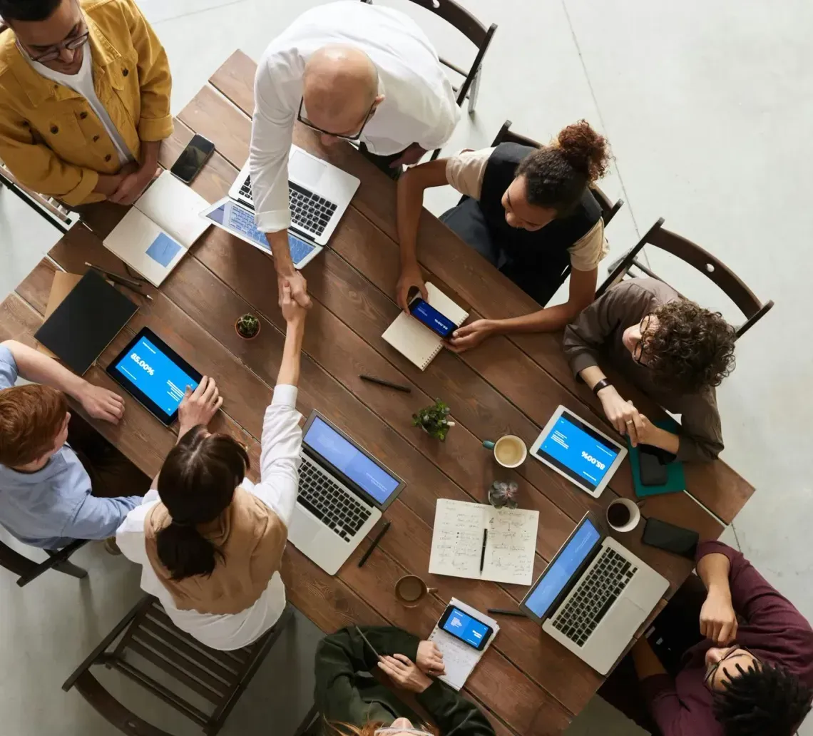 A top-down view of people seated around a wooden table with laptops and tablets, shaking hands in a business meeting.