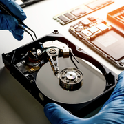 Hands in blue nitrile gloves use tweezers to repair the internal components of an open hard drive on a workbench.