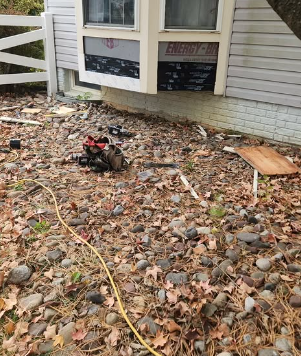 Exterior view of a house with exposed insulation around a window. Debris and leaves cover the ground.