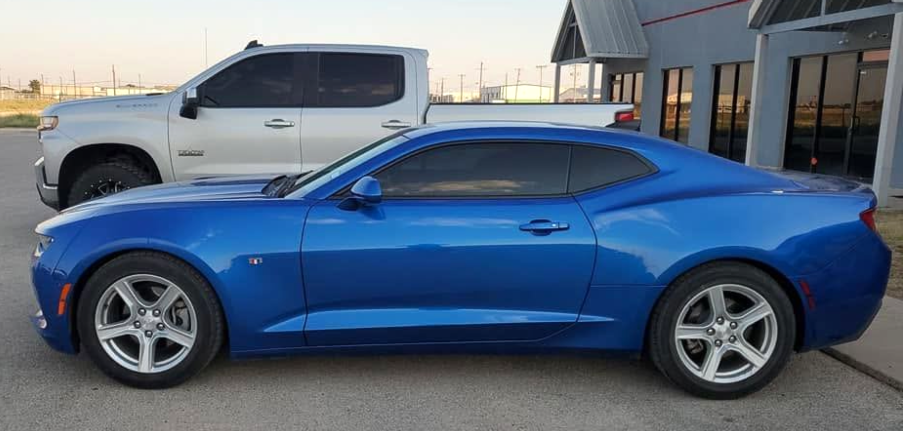 Blue sports coupe parked beside a white pickup truck outside a building