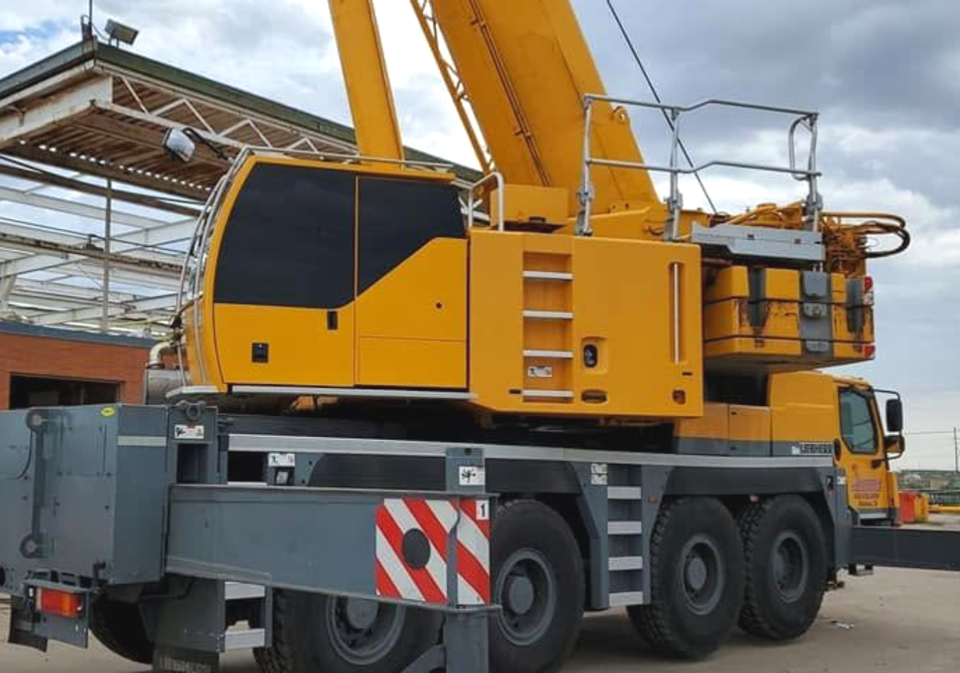 Yellow mobile crane truck parked in an industrial yard, with orange cab and extended boom.