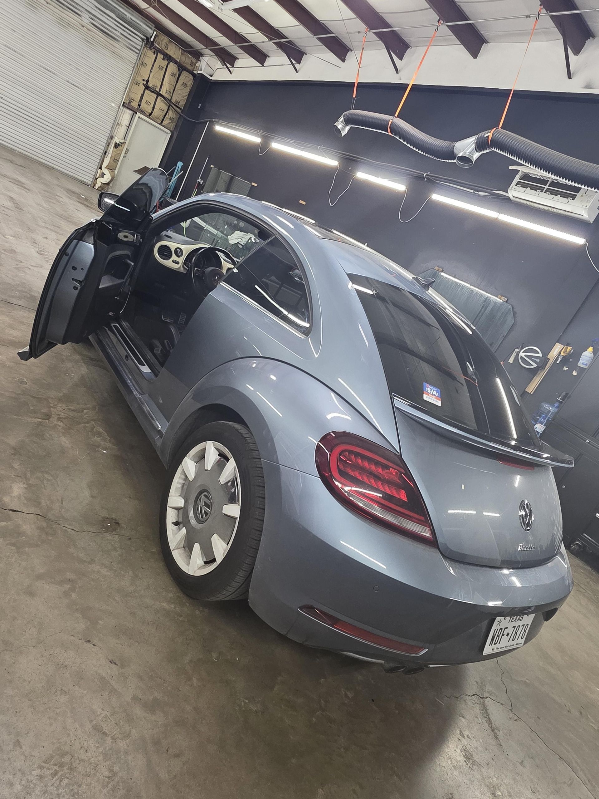 Gray Volkswagen Beetle in a garage, rear view with driver door open and taillights visible.