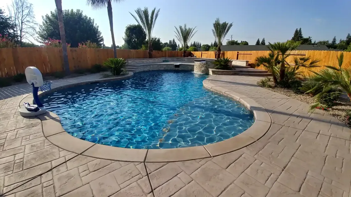 Backyard swimming pool with blue water, tan paver deck, and desert landscaping under clear sky