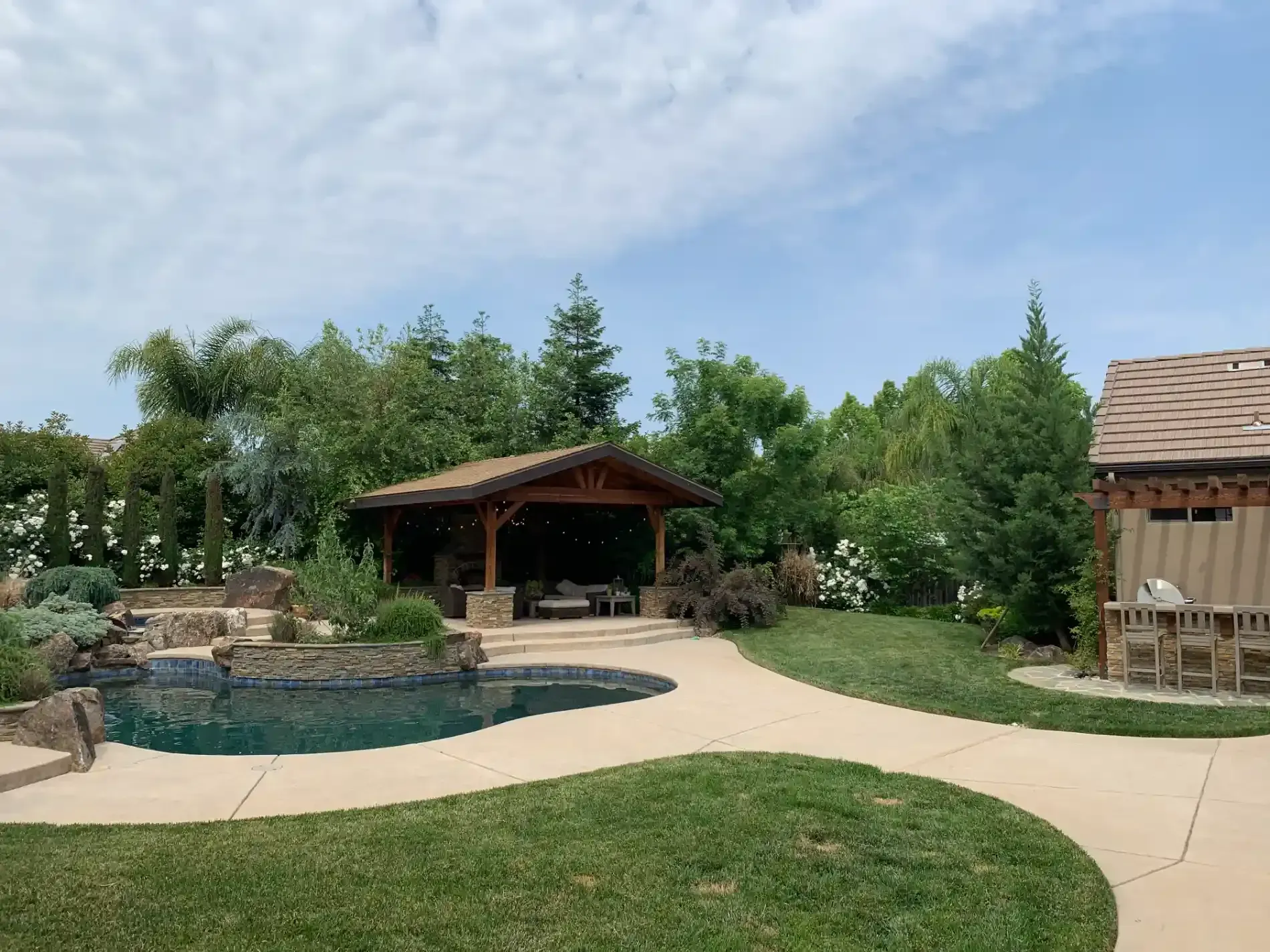Backyard patio with pool, gazebo, curved walkway, and lush landscaping under a blue sky