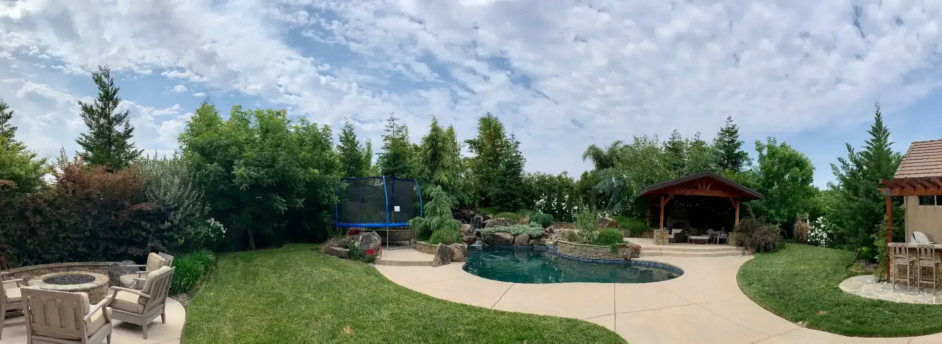 Backyard with curved pool, patio seating, playset, and garden gazebo under trees