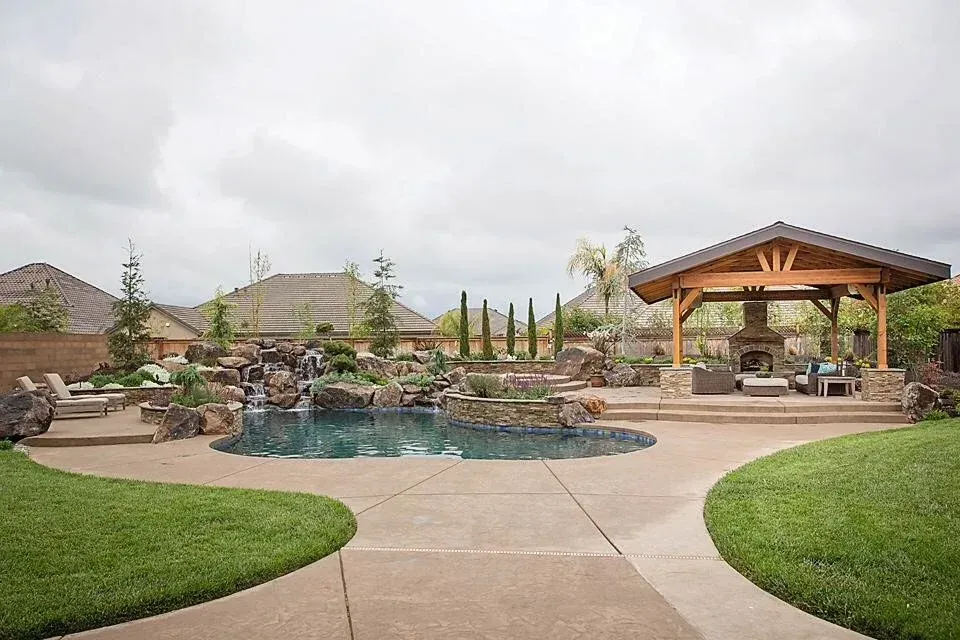 Backyard pool with rock waterfall, patio, and wooden gazebo under overcast sky