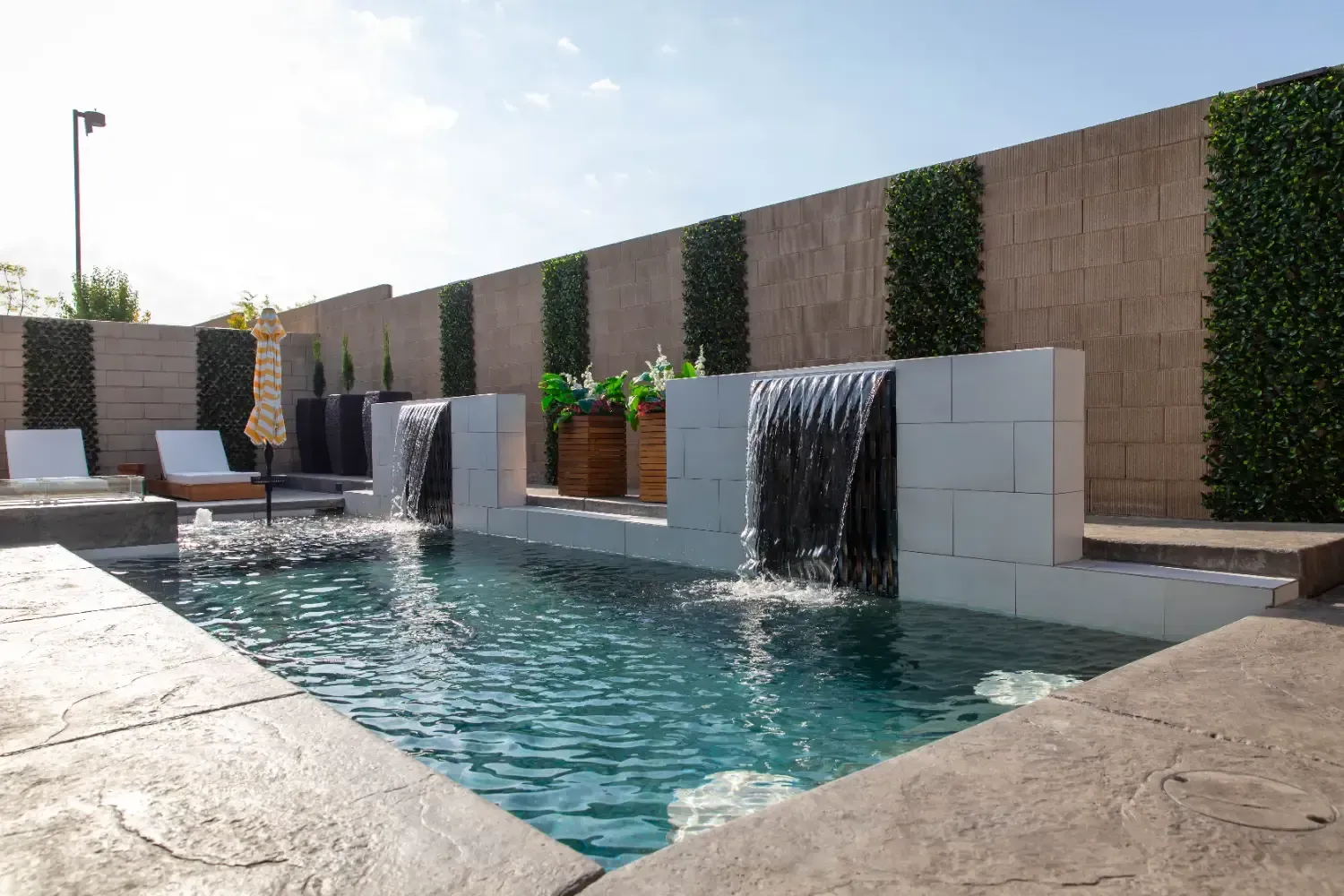 Modern outdoor pool with stone deck, tiled water walls, and green privacy fencing at sunset