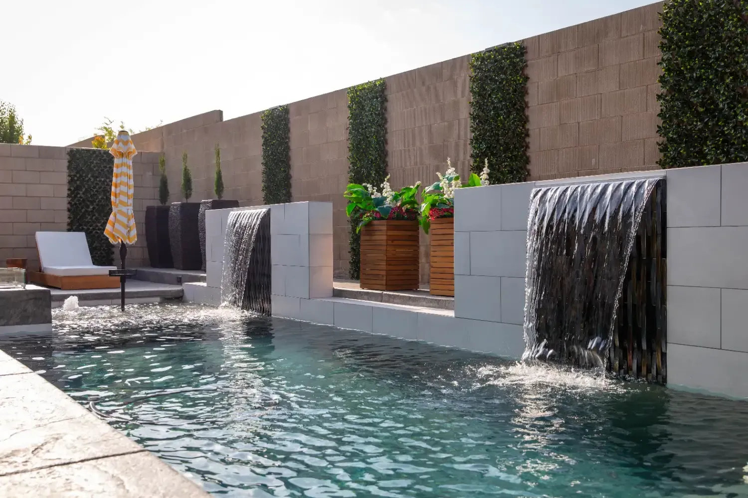 Modern backyard pool with two small cascading waterfalls and lounge chairs along a stone wall
