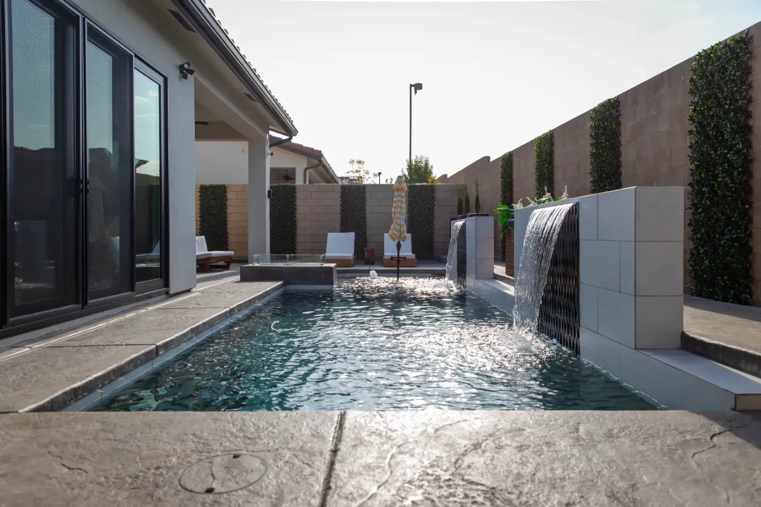Modern backyard pool with waterfalls beside a glass-walled house at sunset