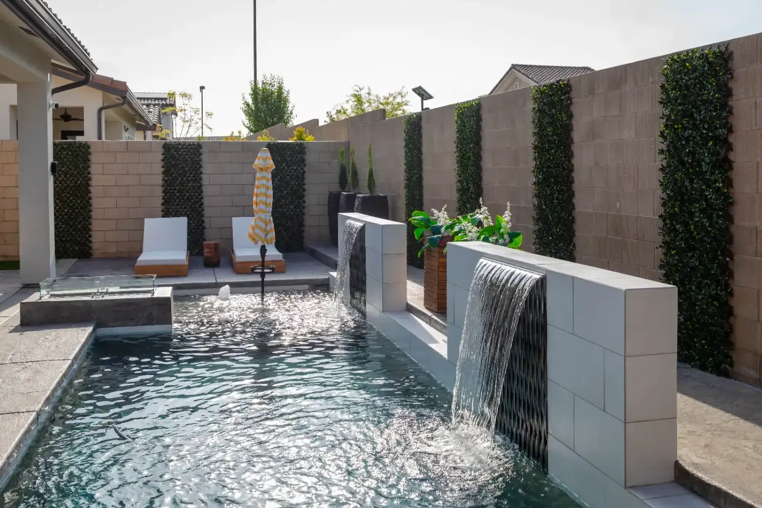 Modern backyard pool with stone waterfall features, lounge chairs, and a beige privacy wall