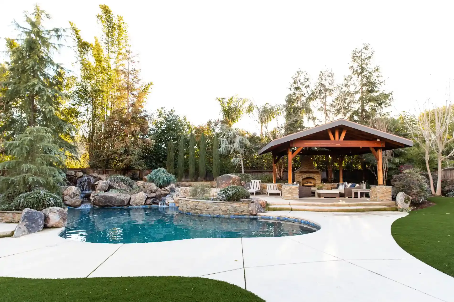 Backyard pool with stone waterfall, wood pavilion, and curved white patio surrounded by trees