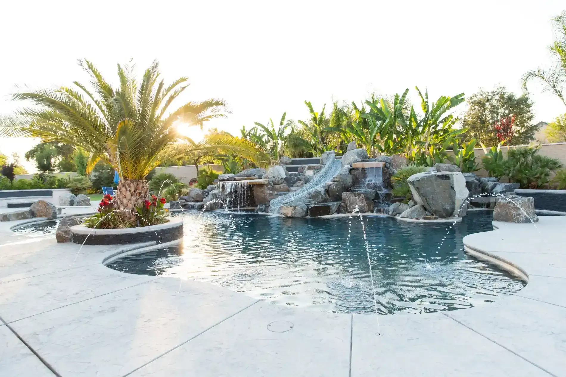 Backyard pool with rock waterfall, palm trees, and a curved stone patio at sunset