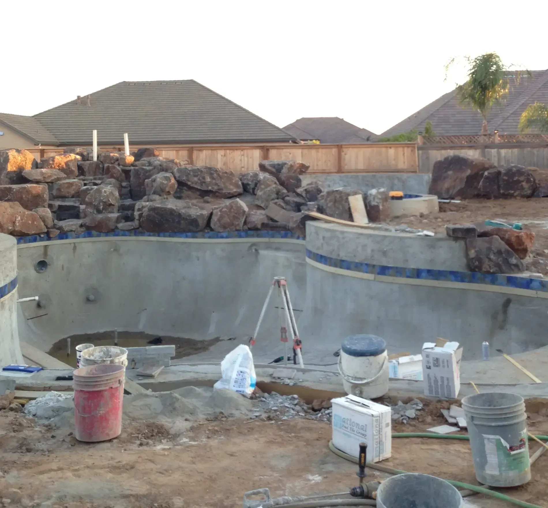 Empty concrete pool under construction with tools, buckets, and rock landscaping in a backyard