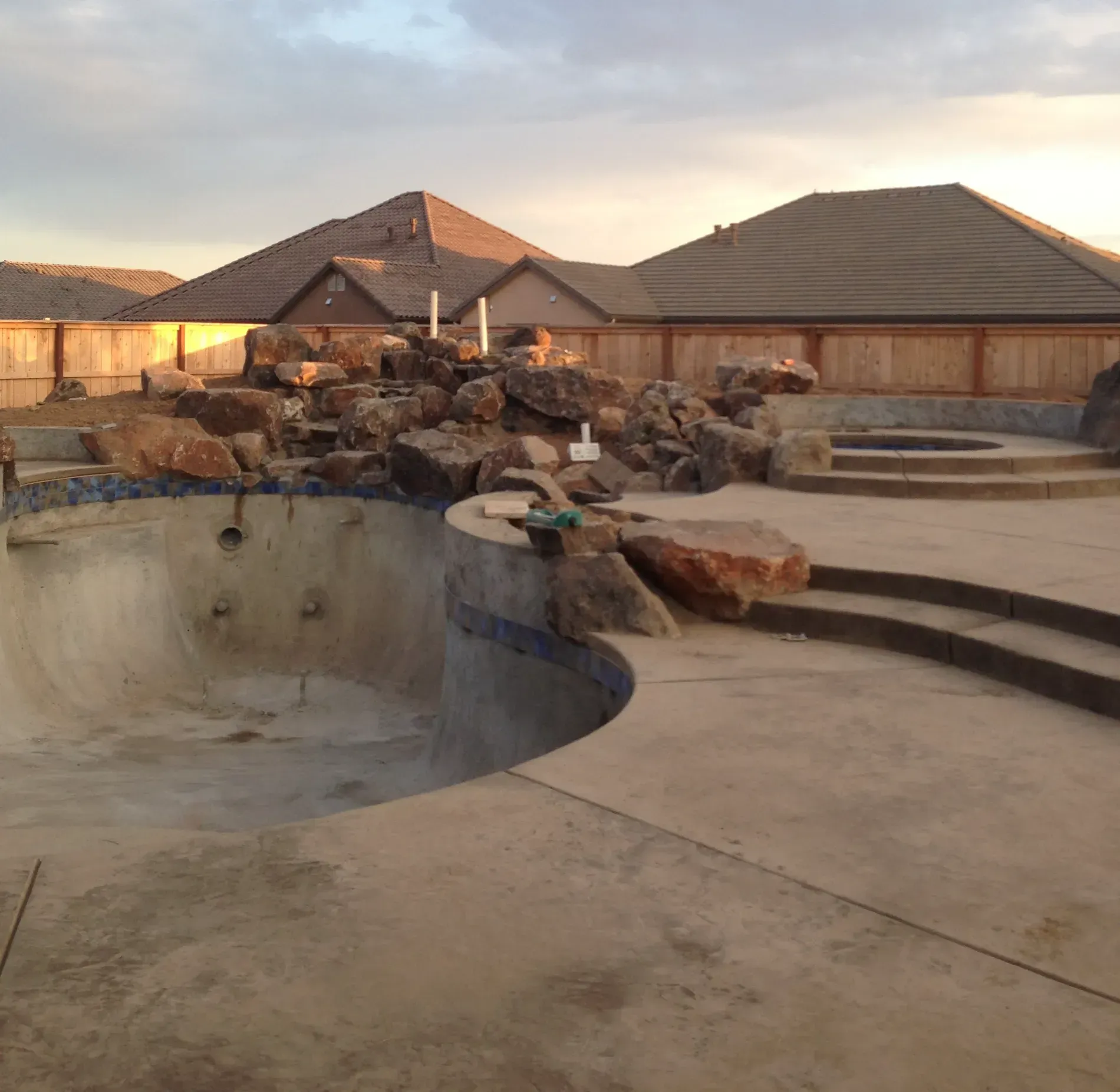 Empty concrete swimming pool beside a stone waterfall and stepped patio at sunset