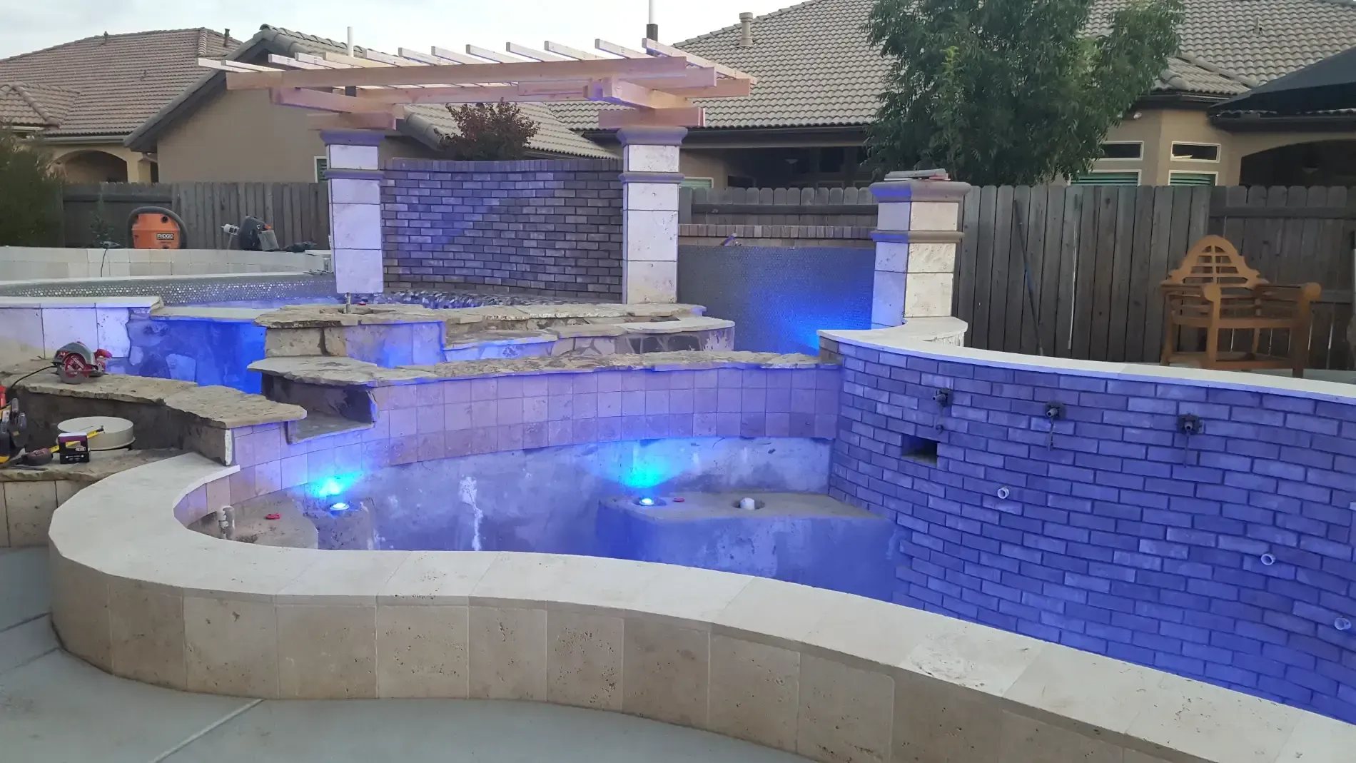 Backyard pool with a curved stone wall, blue LED lighting, and a waterfall feature at dusk