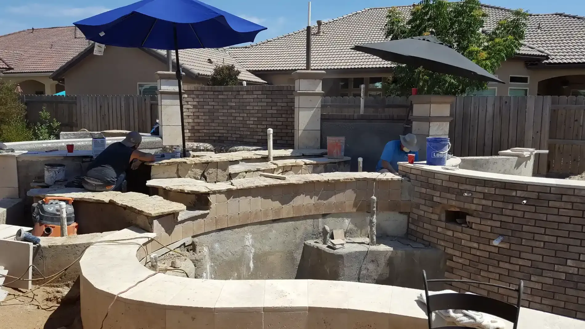 Backyard construction site with a partially built brick pool and patio under blue umbrellas