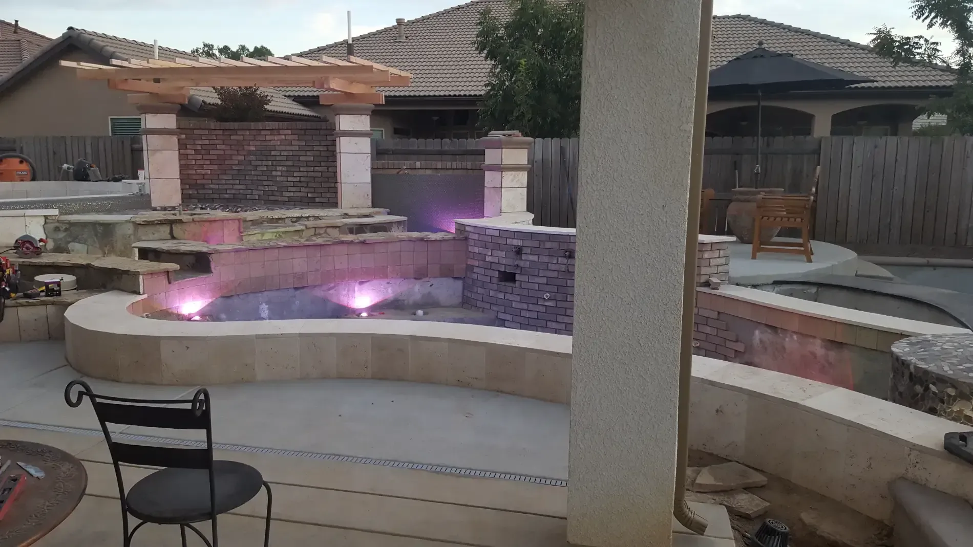 Backyard patio with curved stone fountain lit by purple lights, surrounded by seating and a pergola.