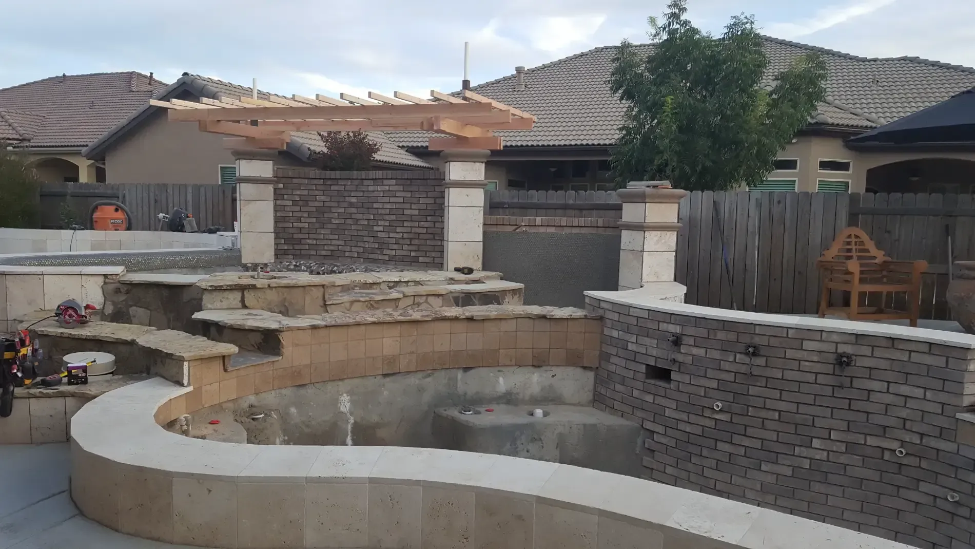 Backyard pool with curved stone walls, patio seating, and pergola in a suburban home setting