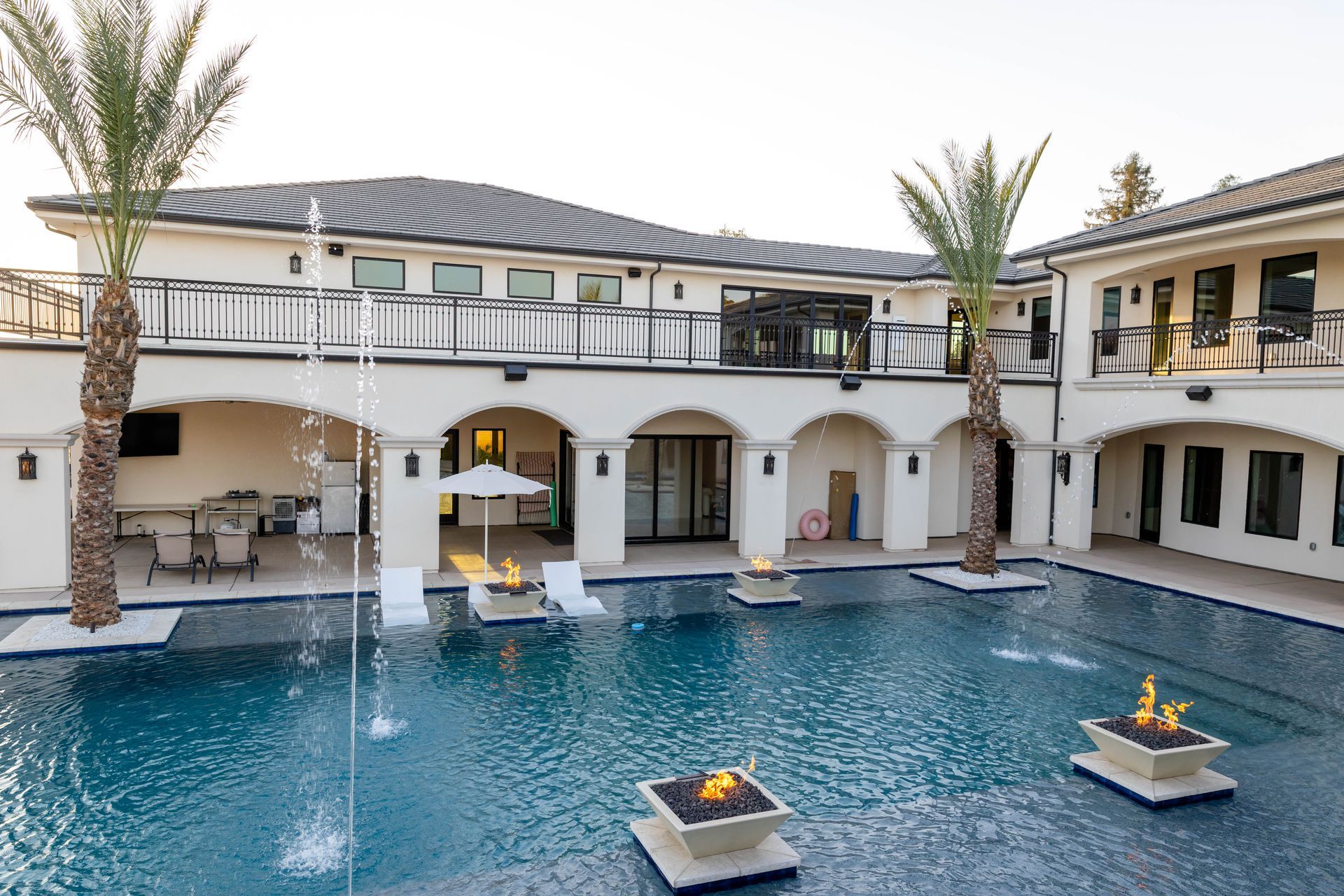 Luxury courtyard with a blue pool, stone fountains, palm trees, and a two-story Mediterranean-style building