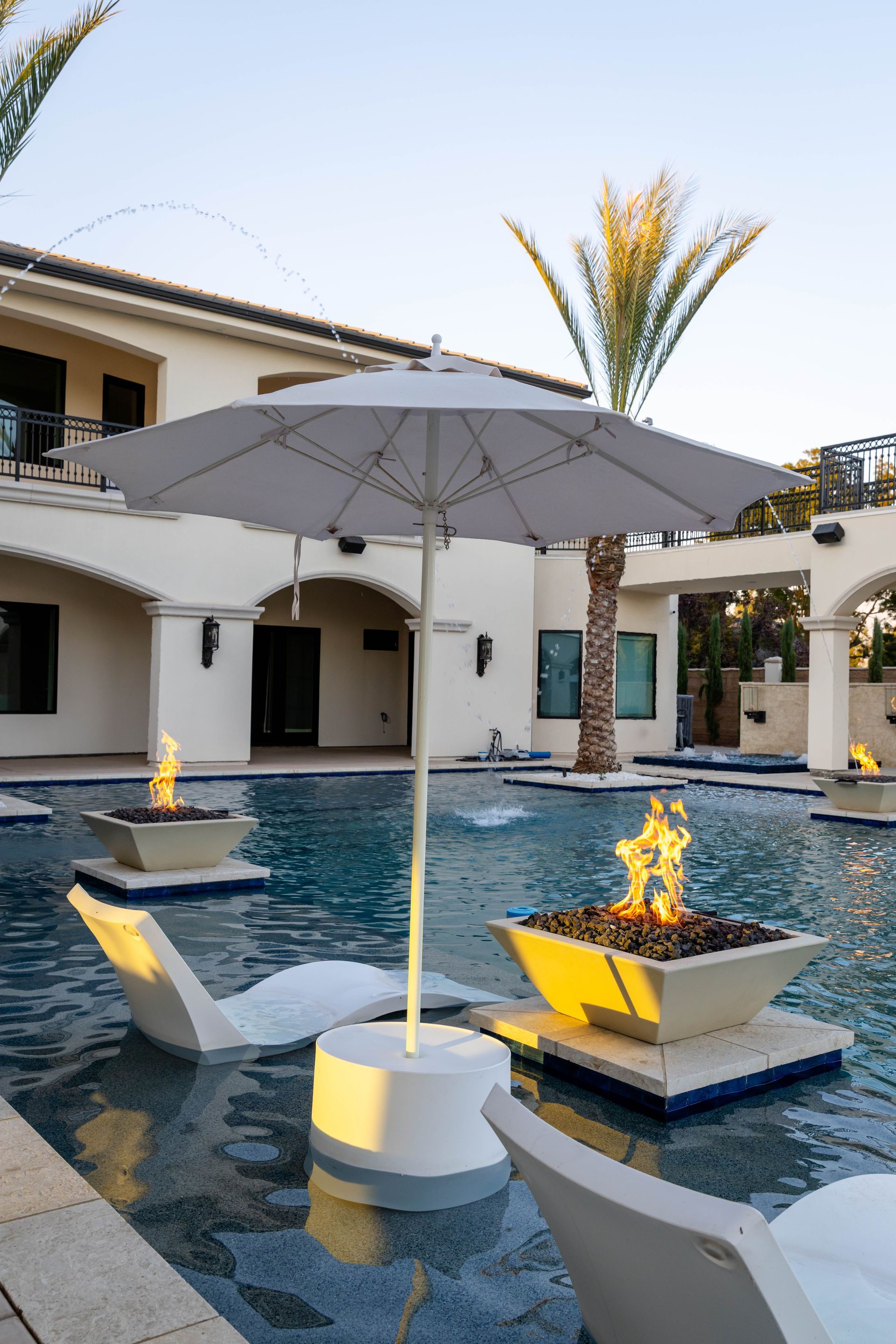 Modern poolside patio with umbrella, lounge chairs, and lit fire bowls around a swimming pool