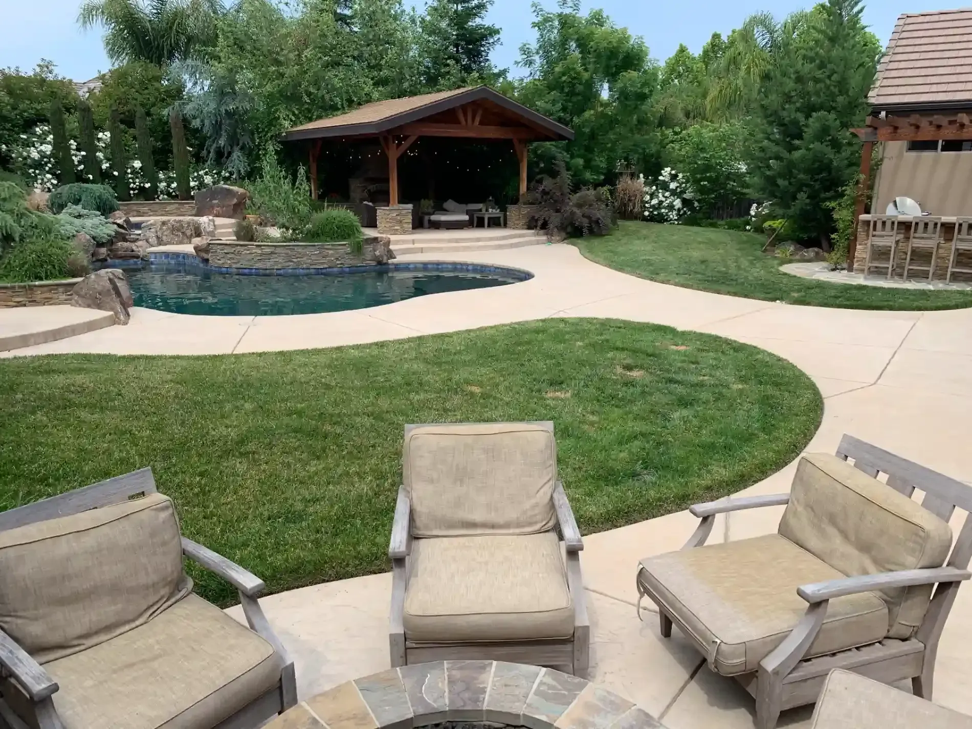Backyard patio with pool, gazebo, lawn, and three beige chairs in the foreground