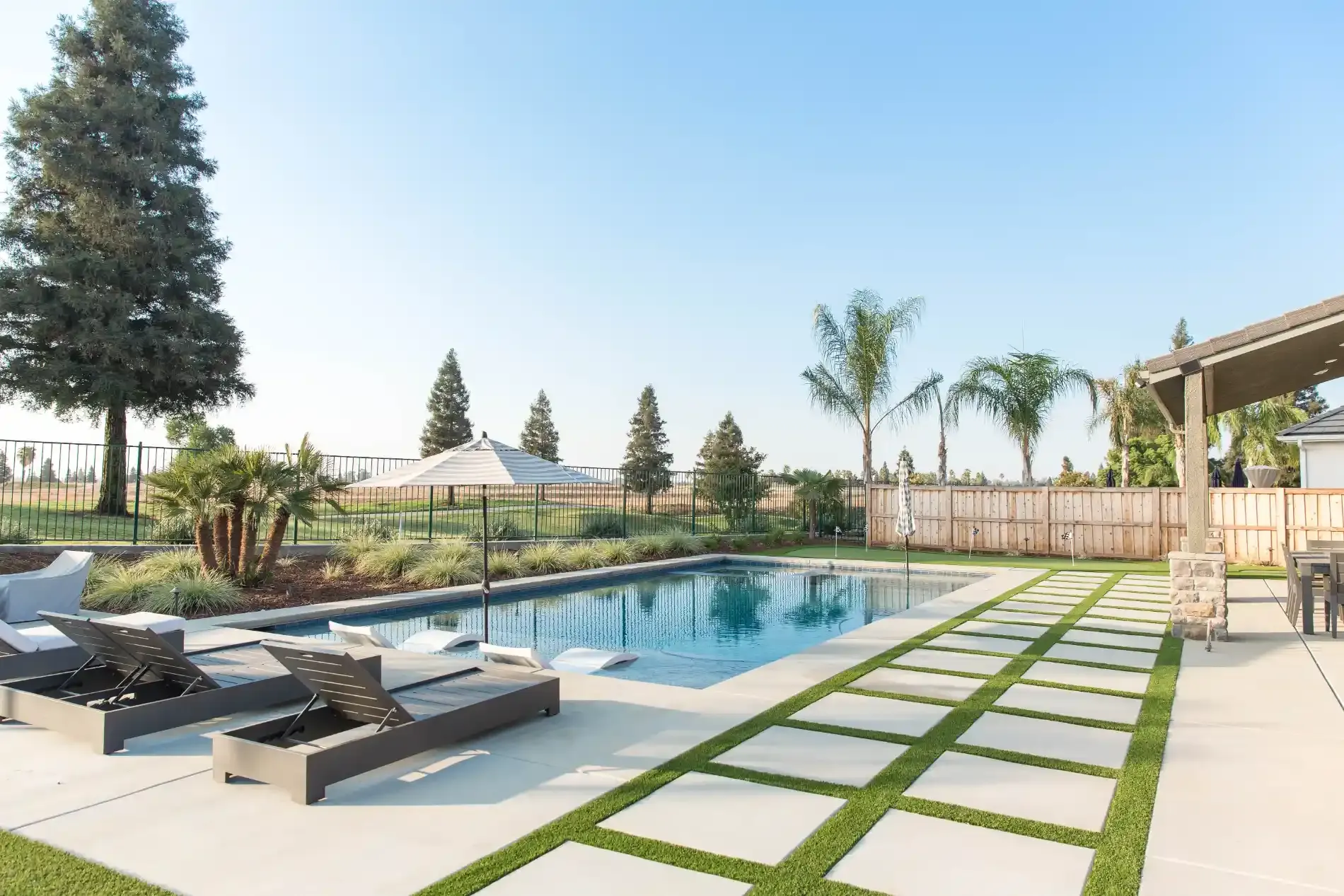 Modern backyard pool with lounge chairs, palm trees, and a tiled patio under a bright sky