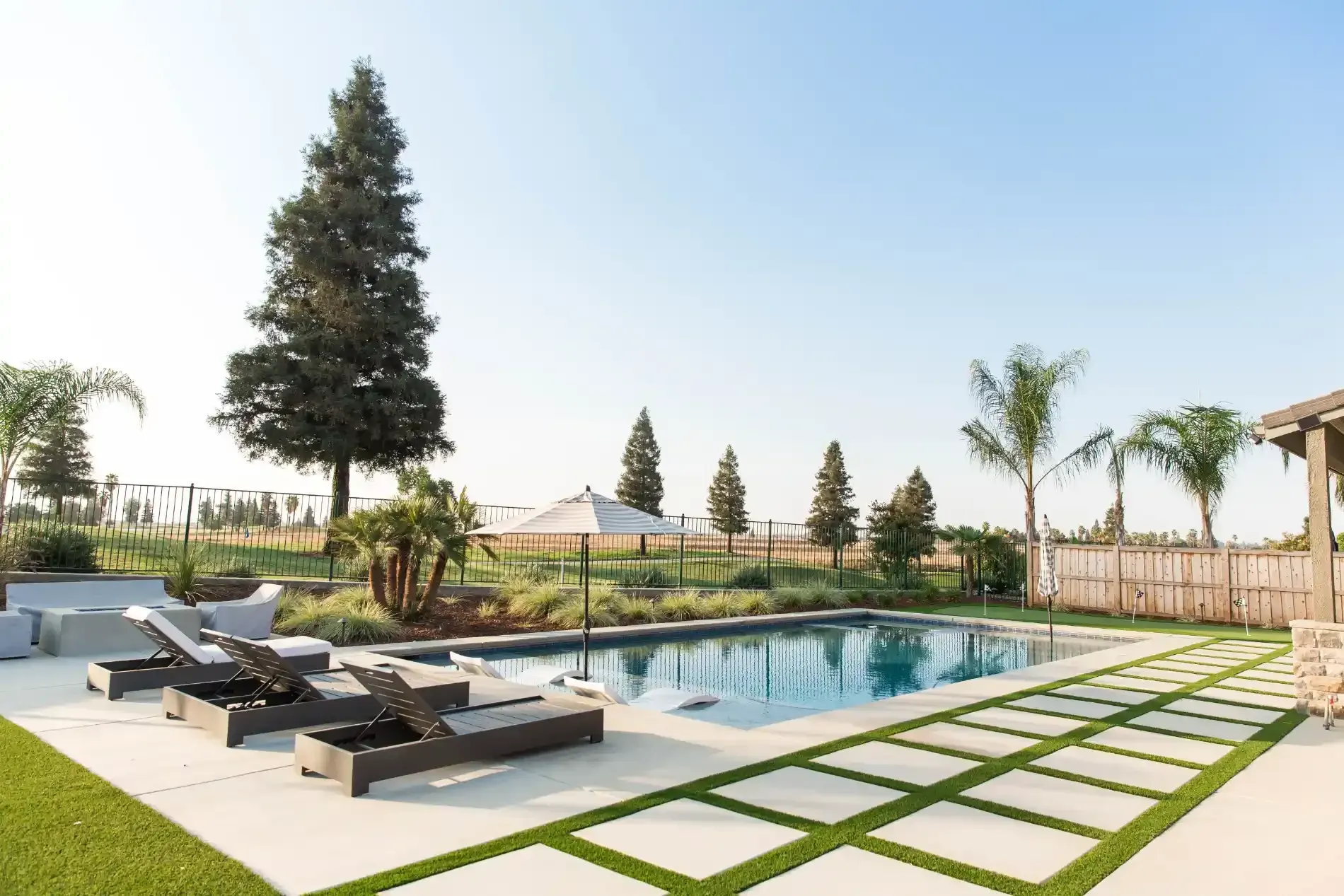 Modern backyard with a swimming pool, lounge chairs, palm trees, and a tiled patio in sunlight