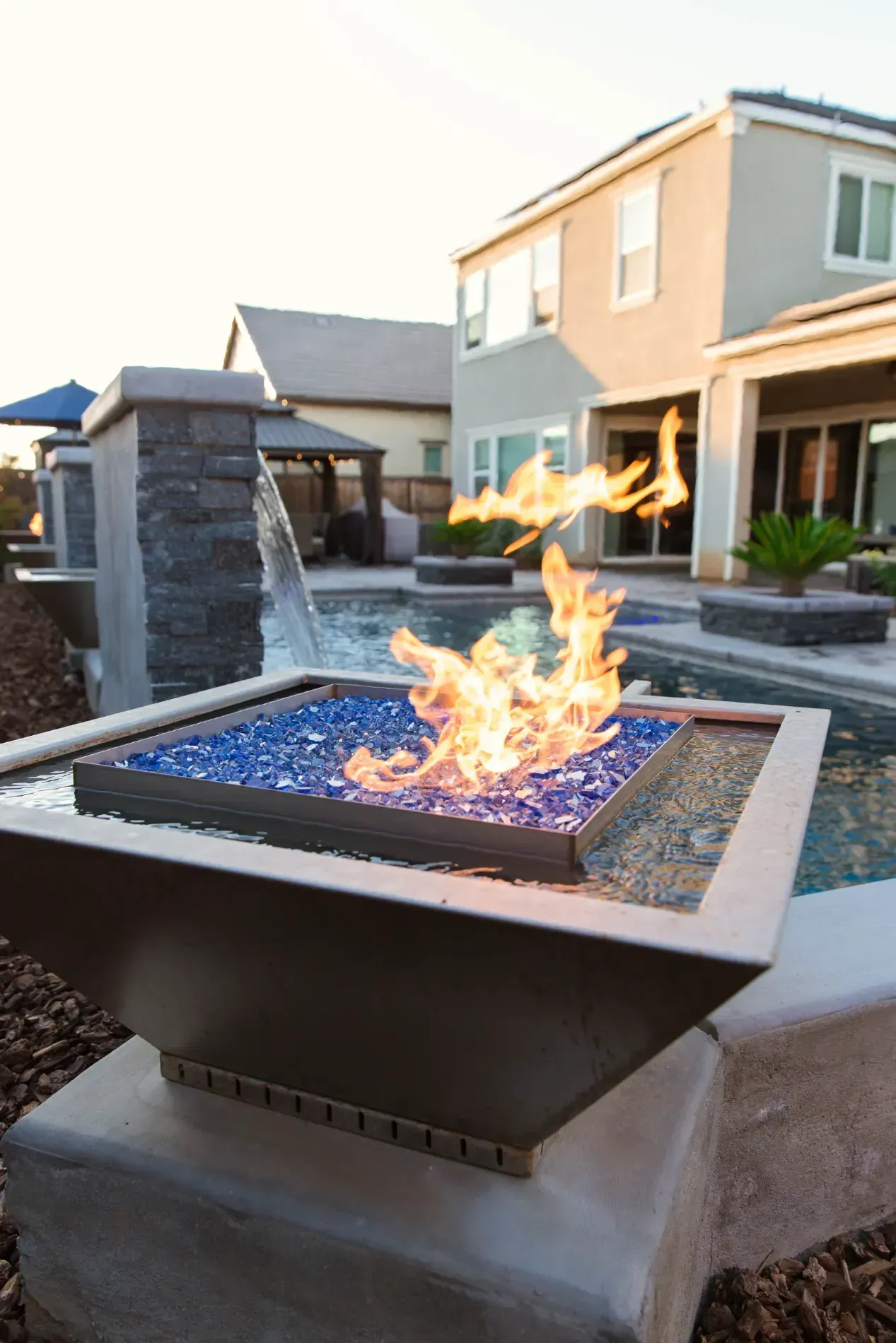 Modern backyard fire pit with flames beside a house and water feature